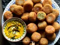 Falafel ricetta originale: croccanti palline dorate, una con un morso rivela il verde interno, accompagnate da una salsa gialla con semi di sesamo nero.