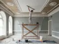 Un operaio sta intonacando il soffitto di una stanza elegante, con impalcature e teli protettivi.