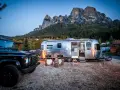Ein Airstream-Wohnwagen und ein schwarzer Geländewagen stehen auf einem **Campingplatz Dolomiten** mit majestätischen Bergen im Hintergrund. Gemütliche Stühle und Lichter schaffen eine einladende Atmosphäre.
