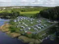 Full campingplats med husbilar och husvagnar vid en sjö, med grönskande träd och bebyggelse i bakgrunden. En idyllisk plats för camping i Alingsås.