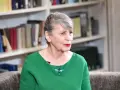 Joanna Wnuk-Nazarowa w zielonej bluzce z broszką w kształcie ważki, na tle regałów z książkami.
