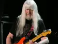 Andy Scott mit seinen charakteristischen langen, weißen Haaren spielt auf einer orangen E-Gitarre.