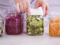 Hände bedecken ein Glas mit fermentiertem Gemüse, Teil einer Auswahl an probiotischen Lebensmitteln in Gläsern.
