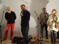 Orkiestra Świętego Mikołaja przygotowuje się do występu. Wokalista z mikrofonem, gitarzysta, skrzypaczka i brodaty mężczyzna w czerwonych spodniach.