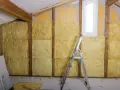 Ocieplenie ścian od wewnątrz wełną mineralną w trakcie prac. Widać drewniane belki, okno i drabinę.