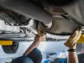 Mani guantate lavorano sul sistema di scarico di un'auto, forse sostituendo il catalizzatore auto.