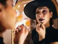 Frau mit Hexenhut und Halloween-Make-up trägt Lippenstift vor einem Spiegel auf.