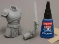 A grey resin knight's torso and arm with a sword are next to a bottle of Loctite Super Glue, suggesting does super glue work on resin for model assembly.