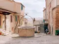Un antico pozzo in mattoni domina una piazzetta di San Giovanni d'Asso, circondata da case in pietra e con dolci colline sullo sfondo.