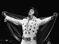 Elvis Presley, der unzählige Tonträger verkaufte, posiert in einem glitzernden weißen Anzug mit ausgebreitetem Umhang.