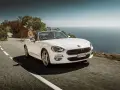 Biały Fiat 124 Spider sunie po krętej drodze nad morzem.