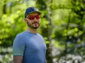 Mężczyzna w czapce i sportowych okularach przeciwsłonecznych, z odbiciem lasu w soczewkach.
