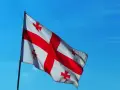 Flaga Gruzji powiewa na tle błękitnego nieba. Białe tło przecina czerwony krzyż, a w każdym z czterech pól znajduje się mniejszy czerwony krzyżyk.