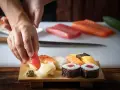 Ręce układają kawałek tuńczyka na ryżu, tworząc sushi. Na desce widać też krewetkę, kalmara, łososia i tamago. To inspiracja, jak zrobić sushi.