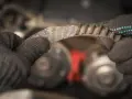Zużyty, piszczący pasek napędowy w rękach mechanika. Widać postrzępione krawędzie i zużyte zęby, co sugeruje potrzebę wymiany.