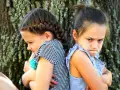 Dwie siostry siedzą plecami do siebie, z ramionami skrzyżowanymi i obrażonymi minami. Może to być odpowiedź na pytanie, dlaczego rodzeństwo się nie lubi.