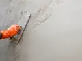 Main d'un ouvrier en gant orange appliquant de l'enduit sur un mur avec une truelle. Apprentissage de comment faire un enduit sur un mur.