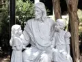 Steinfigur von Jesus, der zwei Kinder umarmt. Er wirkt wie ein liebevoller Gottvater, der seine Schützlinge beschützt.