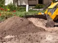 Koparka z łyżką zasypuje ziemię, przygotowując teren pod budowę. W tle kwitnące tulipany.