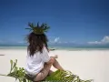 Kobieta w koronie z liści siedzi na plaży, marząc o tym, jak dolecieć na Zanzibar. Błękitne niebo i turkusowa woda zapraszają do podróży.