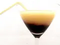 Dwukolorowy drink w kieliszku, z warstwą soku z czarnej porzeczki na dnie i jasnym płynem na górze, ozdobiony żółtą słomką.