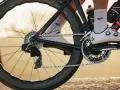 Close-up of a cyclist's shoe and drivetrain, showing the chain, gears, and crankset. The focus is on the power transfer, hinting at the cycling watts being generated.