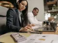 Kobieta w okularach liczy na kalkulatorze, obok stos banknotów i laptop.