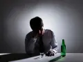 Mężczyzna w białej koszuli i krawacie siedzi przy stole, trzymając się za głowę. Obok stoi szklanka i zielona butelka. To obraz przedawkowania alkoholu.