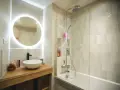 Une **salle de bain blanche** moderne avec un lavabo rond sur un meuble en bois, un miroir lumineux et une douche avec carrelage clair.