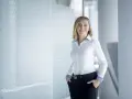 Uśmiechnięta kobieta w białej koszuli i czarnych spodniach z ozdobnym paskiem, stojąca w nowoczesnym wnętrzu.