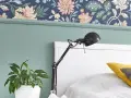 Zielona lamperia pod kwiecistną tapetą, obok łóżka stoi lampka i doniczka z rośliną.