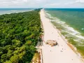 Majówka nad morzem: zielony las, piaszczysta plaża z kilkoma osobami i bunkrem w kamuflażu, szmaragdowe fale Bałtyku.