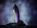 Elegancki perfumy bucik w kolorze fioletowym, ozdobiony złotymi detalami i brokatem, stoi na czarnym postumencie.