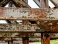Metalowe belki konstrukcji z widoczną łuszczącą się farbą i rdzą. Postępująca korozja niszczy stal, tworząc rdzawy nalot i ubytki.