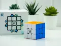 En speed cube 3x3 med orange, vit och blå sida står bredvid sin förpackning. Tre gröna växter i vita krukor syns i bakgrunden.