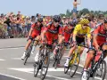 Cyclists in red BMC jerseys race, with one in yellow, as spectators cheer. A perfect scene to watch Tour de France live!