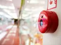 Czerwony przycisk alarmowy, kluczowy element ochrony przeciwpożarowej, zamontowany na jasnej ścianie. W tle rozmyty korytarz centrum handlowego.