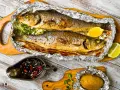 Due branzini cotti al cartoccio, dorati e succulenti, serviti con spicchi di limone, patata al cartoccio e salsa piccante.
