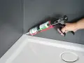 Dłoń trzyma pistolet z tubą **sylikonu**, aplikując go w rogu brodzika prysznicowego.