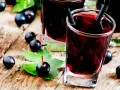 Gläser mit dunklem Johannisbeeren Schnaps, umgeben von frischen Beeren und Blättern auf Holz.