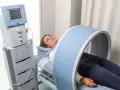 Kobieta leży na łóżku rehabilitacyjnym, poddając się zabiegowi z wykorzystaniem urządzenia generującego pole magnetyczne, które obejmuje jej kolano.