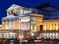 Teatr Wielki w Warszawie, najstarszy teatr w Polsce, nocą. Monumentalna fasada z kolumnami i rzeźbami, oświetlona ciepłym światłem, z samochodami w ruchu ulicznym przed budynkiem.