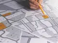 Rysunek geodezyjny z zaznaczonymi działkami, ręka z ołówkiem planuje scalenie gruntów.
