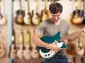 Uśmiechnięty mężczyzna testuje gitarę elektryczną w sklepie muzycznym. W tle widać wiele innych instrumentów.