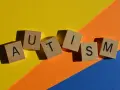 Lettere di legno formano la parola AUTISM su uno sfondo colorato. La domanda "autistici si nasce o si diventa" risuona.