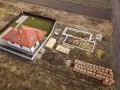 Zabudowa zagrodowa w budowie: gotowy dom z czerwonym dachem, fundamenty, drewno i palety cegieł na placu budowy.