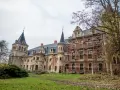 Pałac w Krowiarkach, majestatyczna budowla z wieżami i ceglaną fasadą, otoczony jesienną zielenią, częściowo w rusztowaniach.