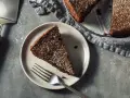 Una fetta di torta all'acqua al cacao, spolverata di zucchero a velo, su un piatto con una forchetta. Accanto, la torta intera e una paletta.