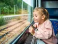 Uśmiechnięta dziewczynka patrzy przez okno pociągu. Zastanawia się, od ilu lat można jeździć samemu pociągiem, marząc o podróżach.