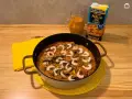 Delicioso arroz a la marinera en paellera, con gambas y mejillones. Al lado, un cartón de caldo para paella de marisco y una jarra con líquido naranja.